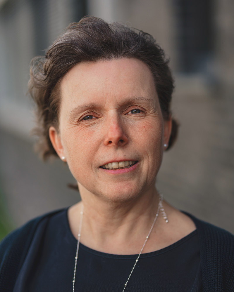 Close-up portrait of a middle-aged woman with short brown hair, wearing a dark top and a delicate necklace, outdoors near a brick wall.